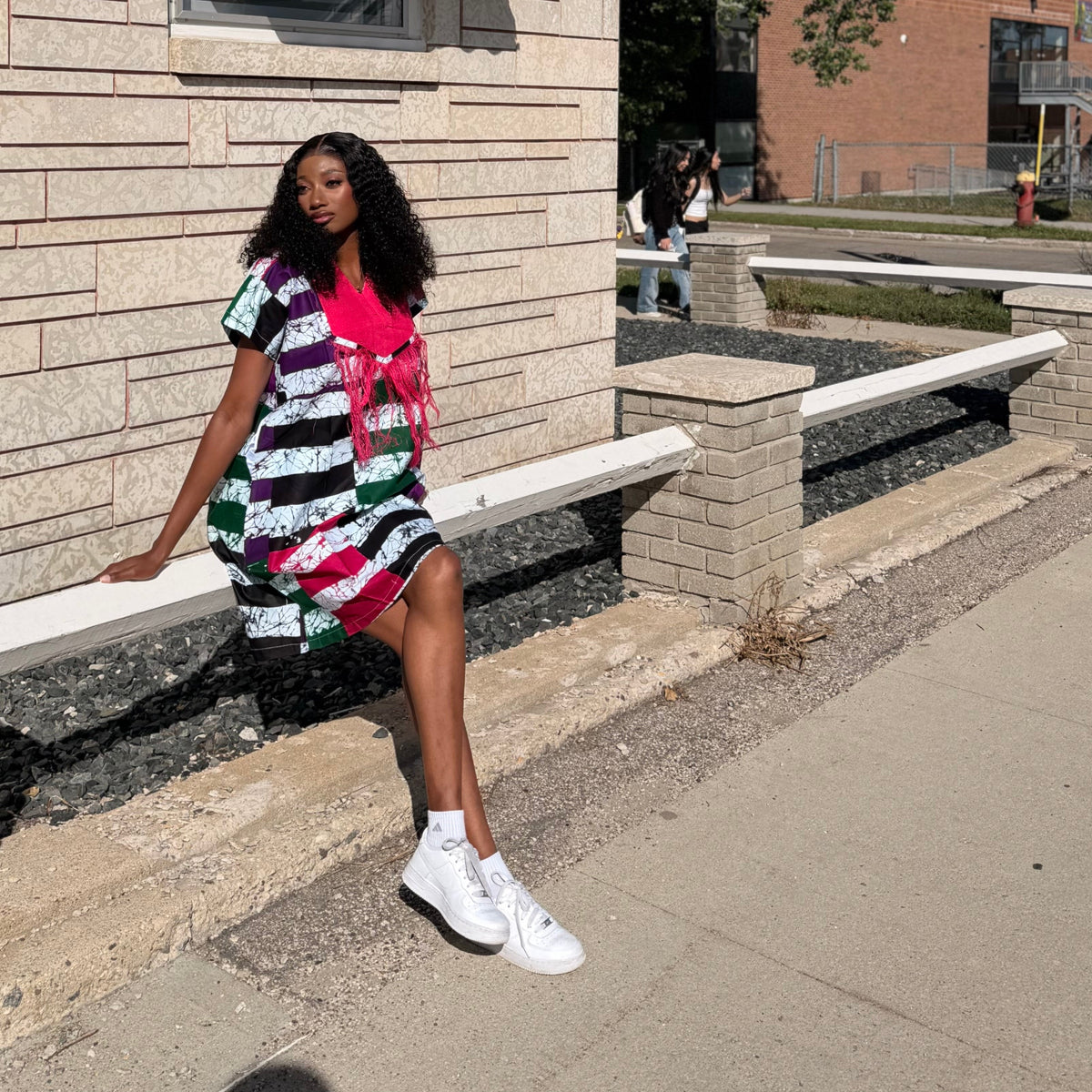 A woman wearing a colorful striped t-shirt dress with a bright pink collar sits on a concrete ledge outside a beige brick building on a sunny day.